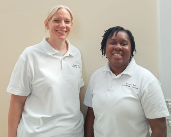 Two smiling speech and language therapists stand together indoors wearing white polo shirts with "Speech and Language Therapy" embroidered on them. They are pictured as part of a Family Hubs initiative providing early speech, language and communication support for children and their families in community settings.